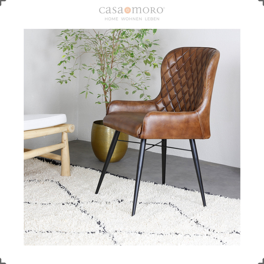 Antik brun läderstol med diamantquiltning på en ljus matta, bredvid en gyllene vas med en grön växt - modern boendeatmosfär med en naturlig touch.