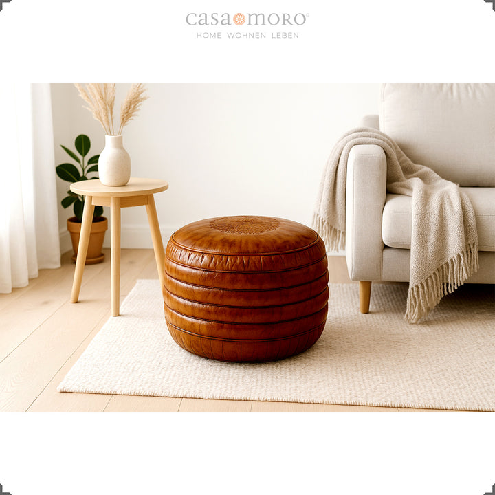 Ljust vardagsrum i skandinavisk stil med en rund läderpuff i brunt på en matta, en beige soffa och en träpall med en vas - en lugn, naturlig atmosfär.