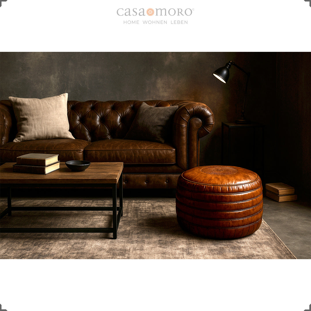 Elegant vardagsrum med Chesterfield-soffa, soffbord och rund läderpuff i brunt - stilren scen i industriell och modern-rustik stil.