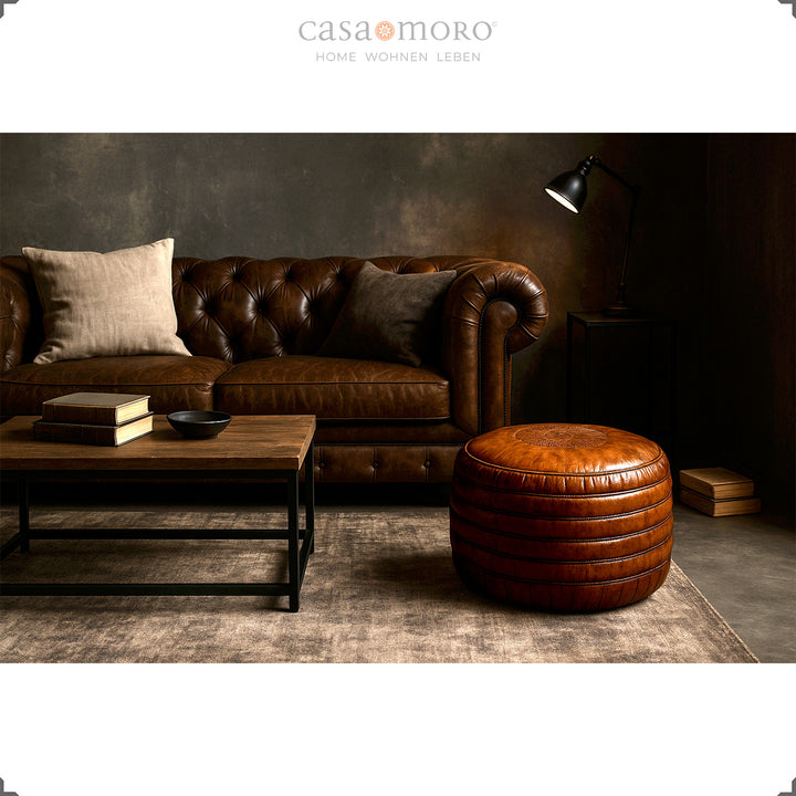 Elegant vardagsrum med Chesterfield-soffa, soffbord och rund läderpuff i brunt - stilren scen i industriell och modern-rustik stil.
