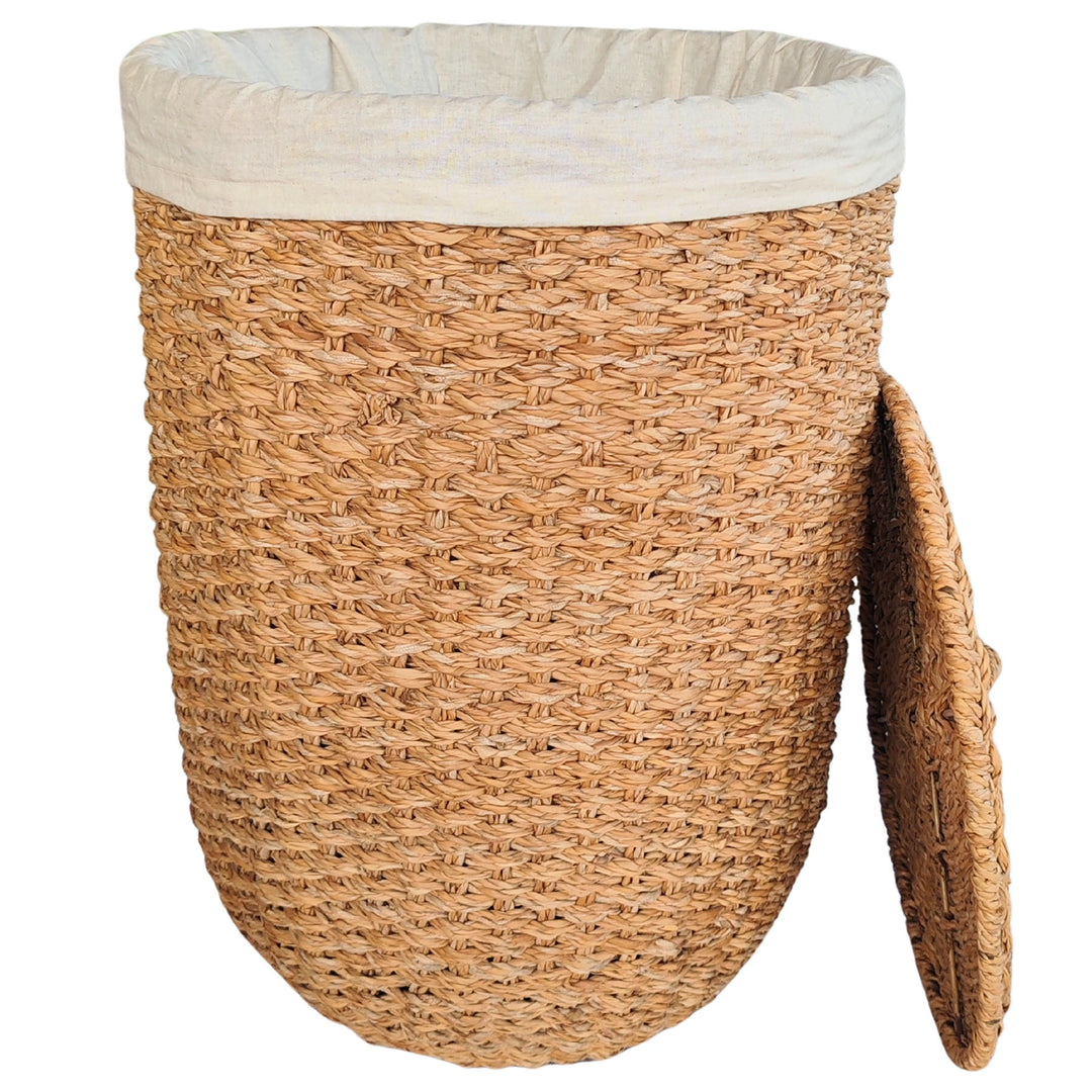 Öppen tvättkorg av naturfärgat sjögräs med en ljus tyginsats och ett lock som vilar mot sidan.